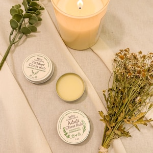 May include: A lit candle in a beige glass, two small tins of chest rub, and a bundle of dried flowers are arranged on a cream-colored cloth. One tin is open, revealing a yellow balm. The tins are labeled "Toddler Chest Rub" and "Adult Chest Rub."