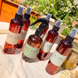 May include: Several amber-colored spray bottles with black spray nozzles are arranged on a white surface. The bottles have labels that read "BUG OFF botanical spray." Green foliage and a wooden fence are in the background. A jar of liquid is also visible.
