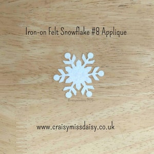 May include: A white iron-on felt snowflake applique. The snowflake has a detailed design with six symmetrical arms and is set against a light brown wooden background. Text at the top reads "Iron-on Felt Snowflake #8 Applique".