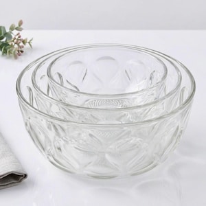 May include: A set of three clear glass mixing bowls nested together. The bowls have a decorative pattern and are arranged on a white surface. A sprig of greenery and a folded linen napkin are in the background.