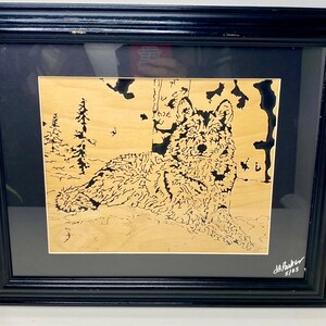 May include: Framed wood art featuring a detailed black and tan wolf resting near a tree. The artwork is set against a light wood background and framed in a black frame. Signed by the artist, J. Parker, 5/05.