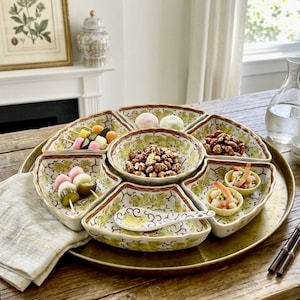 May include: A decorative serving tray with multiple compartments, filled with various snacks. The tray is gold-toned, and the compartments are decorated with a yellow floral pattern. The table setting includes a water carafe, small cups, and chopsticks.