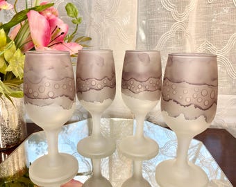 Könnte beinhalten: Vier mattierte Weingläser mit einem dekorativen Design. Die Gläser haben einen weißen Sockel und ein hellviolettes Design mit einem wellenförmigen Muster und kreisförmigen Akzenten. Die Gläser werden auf einer verspiegelten Oberfläche präsentiert.