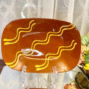 May include: A rectangular brown ceramic serving dish with yellow wavy lines. The dish is displayed on a clear stand, showcasing its glossy finish. The design adds a decorative touch to the tableware.