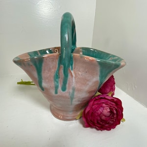 May include: A ceramic basket with a handle, featuring a pink and teal glaze. The basket has a flared shape and is decorated with teal drips. A cluster of deep pink flowers rests beside the basket.