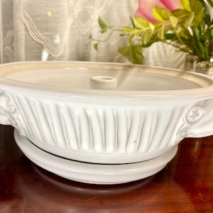 May include: White ceramic casserole dish with a lid. The dish has a ribbed design on the sides and decorative handles. It sits on a matching white base. The dish is likely used for baking and serving.