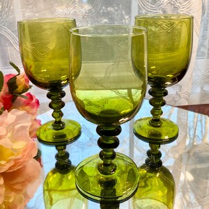 May include: Three green-tinted wine glasses with a decorative pattern on the bowl. The glasses have a round base and a stem with a series of rounded shapes. The glasses are arranged on a reflective surface.