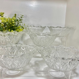 May include: A collection of six clear glass bowls with a decorative, scalloped edge. The bowls are arranged on clear acrylic stands, and each has a detailed, embossed pattern. A small arrangement of green and yellow artificial flowers is in one bowl.
