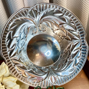 May include: A silver-colored, decorative bowl with a detailed leaf design around the rim and a central, concave area. The bowl is supported by a green, scrolled metal stand, suggesting a holiday or festive theme.