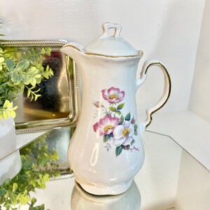 May include: A white ceramic pitcher with a lid, featuring a floral design of pink and white flowers with yellow centers and green leaves. The pitcher has a gold trim on the handle and spout. A vintage-style decorative item.