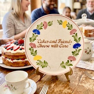 May include: A decorative plate with a floral border in pink, yellow, and blue, and the text "Cakes and Friends Choose with Care." The plate is displayed on a gold-colored stand, with a cake and teacup in the foreground.