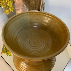 May include: A handmade, ceramic bowl with a pedestal base. The bowl is a warm, earthy brown color with a lighter rim and a spiral pattern in the center. The bowl is likely used for serving or display.