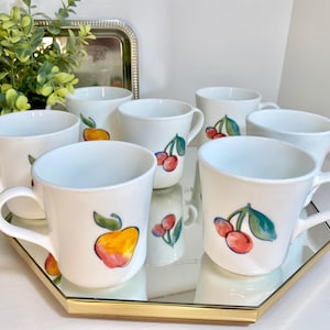 May include: A set of seven white ceramic mugs with fruit illustrations. Each mug features a different fruit design, including apples and cherries, in vibrant colors. The mugs are displayed on a gold-trimmed, mirrored tray.