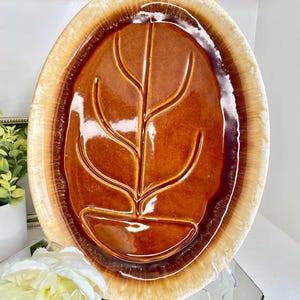 May include: Oval-shaped brown ceramic serving platter with a leaf design. The platter has a glossy finish and a light brown rim. The leaf design is raised and adds texture to the surface. A decorative piece for serving food.