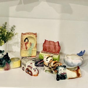 May include: A collection of vintage ceramic planters in various shapes and colors. Includes a bear, a train, a cat, a bird, and a woman. The planters are arranged on a white shelf, with a small plant in a white pot in the background.