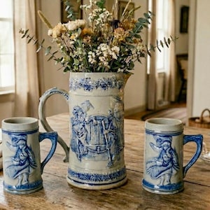 May include: A rustic still life featuring a large ceramic pitcher and two matching mugs, all adorned with blue and white relief designs. The pitcher holds a bouquet of dried flowers. The scene is set on a wooden table.