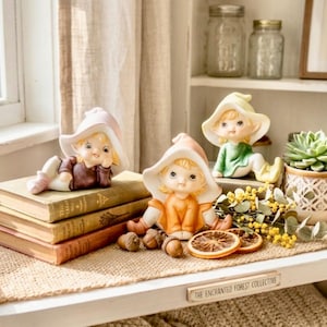 May include: Three ceramic fairy figurines with large hats in pastel colors are displayed on a shelf with books, a succulent plant, and dried oranges. The shelf is decorated with natural elements and a sign that reads "The Enchanted Forest Collective."