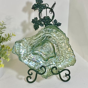 May include: A decorative, iridescent green glass plate with a textured surface, displayed on a dark green metal stand. The stand features a grape vine design at the top. The plate has a unique, organic shape.