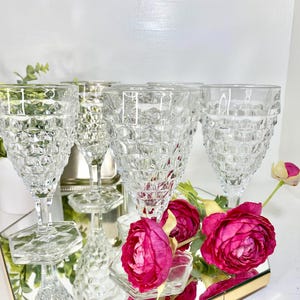 Puede incluir: Un conjunto de copas de vino transparentes con patrones y bases hexagonales. Las copas están dispuestas sobre una superficie espejada, junto con vibrantes rosas rosas y vegetación. Las copas tienen un patrón texturizado en forma de diamante.