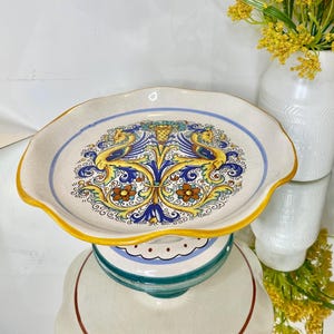 May include: A ceramic pedestal cake stand featuring a scalloped edge with a yellow rim. The flat surface displays a central hand-painted design of two yellow sea creatures with blue wings, surrounded by blue and brown floral motifs on cream.
