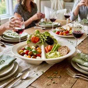 May include: A white divided serving platter filled with colorful cherry tomatoes, carrots, celery, crackers, and dip. The platter is on a wooden table set for a meal, with wine glasses and plates.