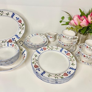 May include: A white porcelain dinnerware set with a floral design in red, green, and blue. The set includes plates, bowls, saucers, and teacups. A serving platter and a creamer are also included. A bouquet of pink flowers is in the background.