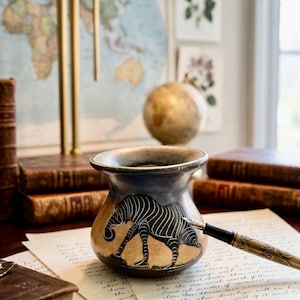 May include: A decorative ceramic vase with a zebra design. The vase is primarily tan with a dark blue rim and features a black and white zebra illustration. A vintage fountain pen rests on a sheet of paper, suggesting a writing theme.