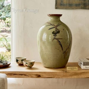 May include: A large, olive green ceramic vase with a brown rim and a painted floral design. Two small, decorative bowls with floral patterns sit on a wooden shelf. The shelf also holds books and a small sign.