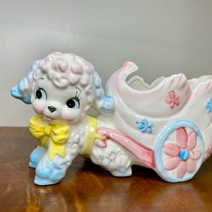 May include: A vintage ceramic poodle planter in pastel colors. The poodle is white with blue and pink accents, pulling a pink and white carriage decorated with flowers. The planter is a charming decorative piece.