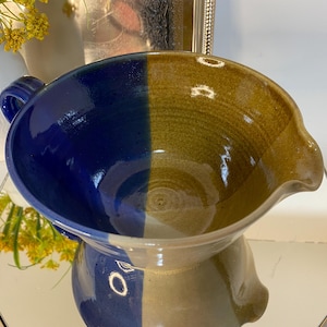 May include: A handmade ceramic bowl with a unique two-tone design. Half of the bowl is a deep blue, while the other half is a warm, earthy brown. The bowl has a spout and a handle, making it ideal for serving or display. The bowl is a decorative kitchenware item.