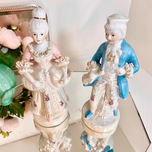 May include: Two porcelain figurines of a man and woman in 18th-century attire. The woman wears a pink dress with floral accents, while the man has a blue coat. Both figures have detailed facial features and are on round bases.