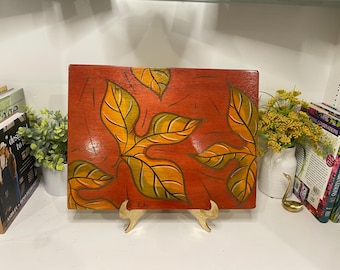 May include: A rectangular wooden art piece featuring a leaf design. The leaves are painted in shades of yellow, orange, and green against a reddish-orange background. The artwork is displayed on a gold-colored stand, showcasing a nature-inspired theme.