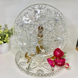 May include: A clear, vintage glass serving platter with a starburst design. The platter is supported by a gold-colored metal stand. Red flowers are placed near the base of the platter. The platter is likely used for serving food or displaying decorative items.
