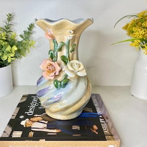 May include: A decorative ceramic vase with a ruffled rim, featuring pastel colors and sculpted roses. The vase has a swirling design and is adorned with pink, white, and green floral accents. It is a charming piece for home decor.