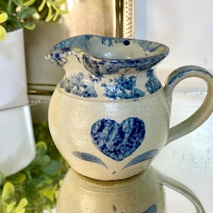 May include: A small, cream-colored ceramic pitcher with a blue heart design and blue accents. The pitcher has a rounded body, a handle, and a spout. The blue heart is centered on the front.