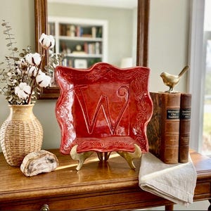May include: A red decorative plate with the letter "W" in the center, displayed on a gold stand. The plate has a decorative border. A vase with cotton stems and a small brass bird are also visible.