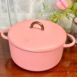 May include: A pink cast iron Dutch oven with a matching lid. The lid has a bronze-colored handle and a decorative emblem. The pot has two side handles for easy carrying. The pot is sitting on a wooden surface.
