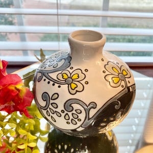Puede incluir: Un peque&ntilde;o jarr&oacute;n redondo de cer&aacute;mica con base blanca, adornado con patrones florales y abstractos en gris, amarillo y negro. El jarr&oacute;n tiene una abertura estrecha y un acabado brillante, adecuado para exhibir flores peque&ntilde;as o como pieza decorativa.