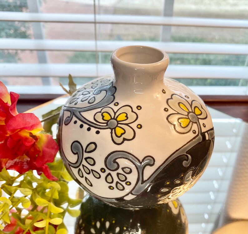 Puede incluir: Un peque&ntilde;o jarr&oacute;n redondo de cer&aacute;mica con base blanca, adornado con patrones florales y abstractos en gris, amarillo y negro. El jarr&oacute;n tiene una abertura estrecha y un acabado brillante, adecuado para exhibir flores peque&ntilde;as o como pieza decorativa.