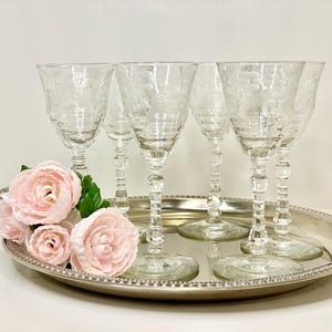 May include: Six clear, etched wine glasses with a floral design, arranged on a silver serving tray. The glasses have a decorative stem. Pink roses and green leaves are placed on the tray. The tray has handles.