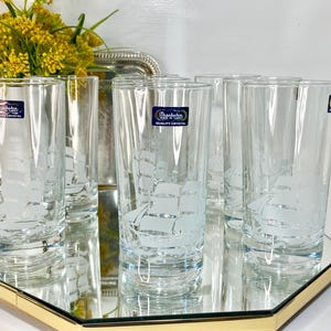 May include: Set of six clear crystal highball glasses. Each glass features an etched sailboat design. The glasses are displayed on a mirrored tray with a gold-colored trim. A bouquet of yellow flowers sits in the background.