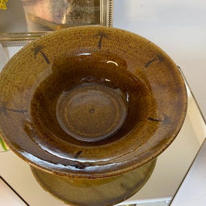 May include: A brown ceramic bowl with a wide rim and a central depression. The bowl has a textured surface and is decorated with dark brown palm tree designs around the rim. The bowl is sitting on a mirrored surface.