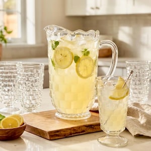 May include: A clear glass pitcher and matching glasses filled with lemonade and ice. The pitcher and glasses have a textured pattern. Slices of lemon and mint leaves are visible in the pitcher and glass. A wooden bowl with lemon slices is also present.