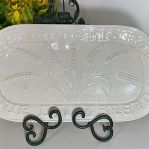 May include: A white rectangular serving platter with embossed wheat stalks and the words "Give Us This Day Our Daily Bread" around the border. The platter is supported by a dark green metal stand.