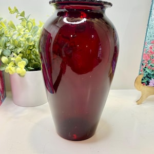 May include: A tall, ruby-red glass vase with a ruffled rim. The vase has a smooth, glossy surface and a slightly flared base. The vase is empty and sits on a white surface. Decorative home decor.