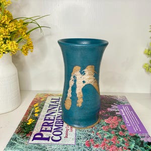May include: A teal ceramic vase with a flared top and a textured, abstract gold design. The vase is displayed on a magazine with the text "PERENNIAL COMBINATIONS". Yellow flowers in a white vase are in the background.