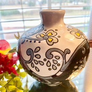 Puede incluir: Un peque&ntilde;o jarr&oacute;n redondo de cer&aacute;mica con cuello gris y cuerpo blanco. Decorado con dise&ntilde;os florales y de volutas en negro, gris y amarillo. El jarr&oacute;n est&aacute; sobre una superficie reflectante.
