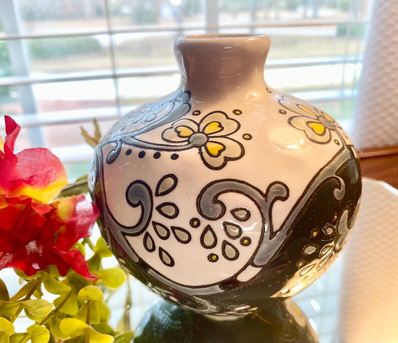 Puede incluir: Un peque&ntilde;o jarr&oacute;n redondo de cer&aacute;mica con cuello gris y cuerpo blanco. Decorado con dise&ntilde;os florales y de volutas en negro, gris y amarillo. El jarr&oacute;n est&aacute; sobre una superficie reflectante.