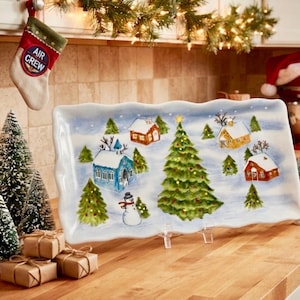 May include: A rectangular ceramic serving platter with a wavy edge, featuring a hand-painted winter village scene. The design includes a large central Christmas tree, several small houses with snow-covered roofs, a snowman, and evergreen trees.