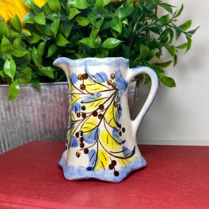 May include: A small, ceramic pitcher with a white base, adorned with yellow and blue leaf and berry designs. The rim and base are trimmed in blue. The pitcher has a handle and a spout. The pitcher is approximately 5 inches tall.